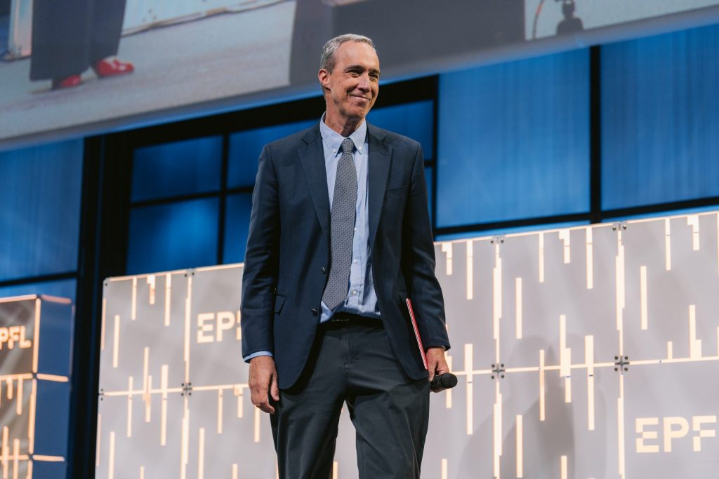 Daniel Huttenlocher on stage at EPFL, in a suit, holding a microphone and a red diploma.