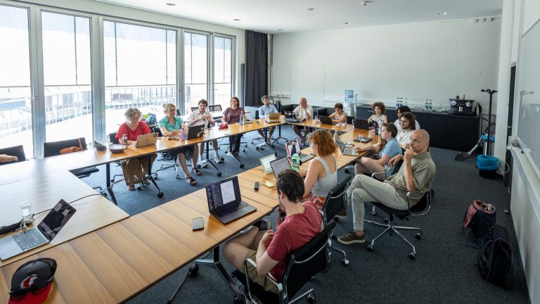 L'Assemblée d'Ecole assiste à une réunion dans une salle lumineuse avec grandes fenêtres. Elles sont assises autour de tables en O, avec ordinateurs, bouteilles d’eau et carnets. L’ambiance est détendue mais concentrée. En arrière-plan : un coin café et un tableau blanc.